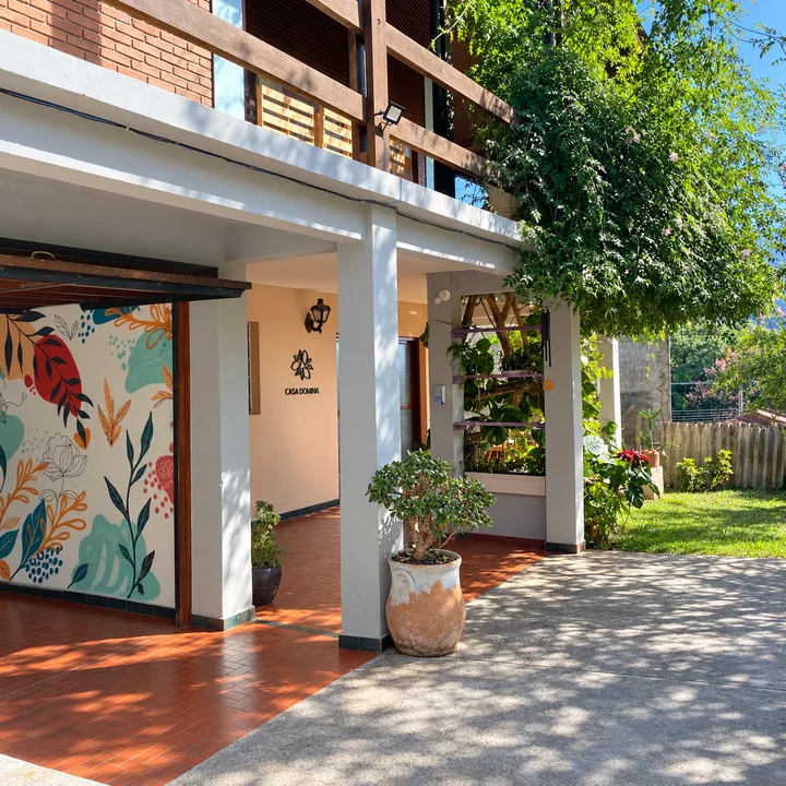 Pátio da Casa Domina em Mairiporã: varanda com mural de folhagens, plantas, jardim e o logo do estúdio